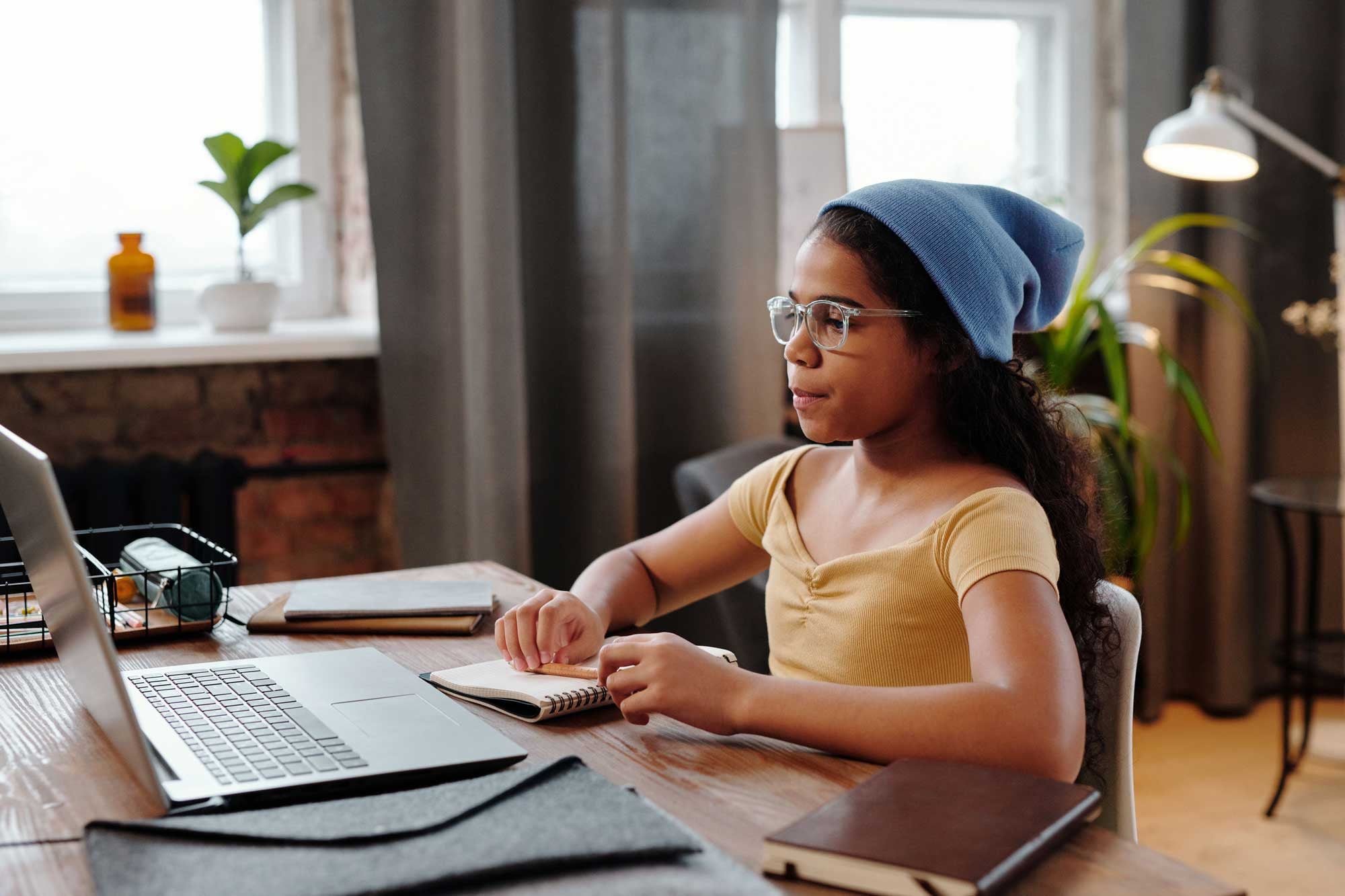 Child in blue beanie and glasses studying at a desk with a laptop and notebook.